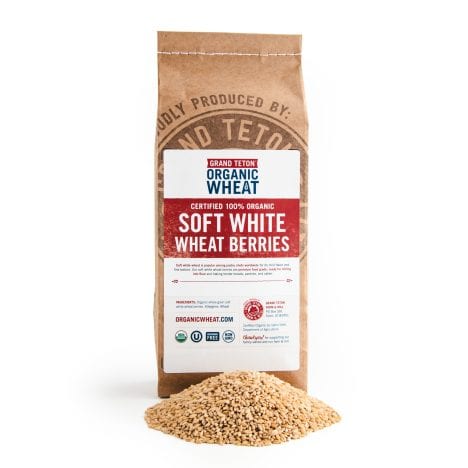 Brown kraft paper bag of Grand Teton Organic Soft White Wheat Berries standing behind a pile of whole golden wheat berries on a white background